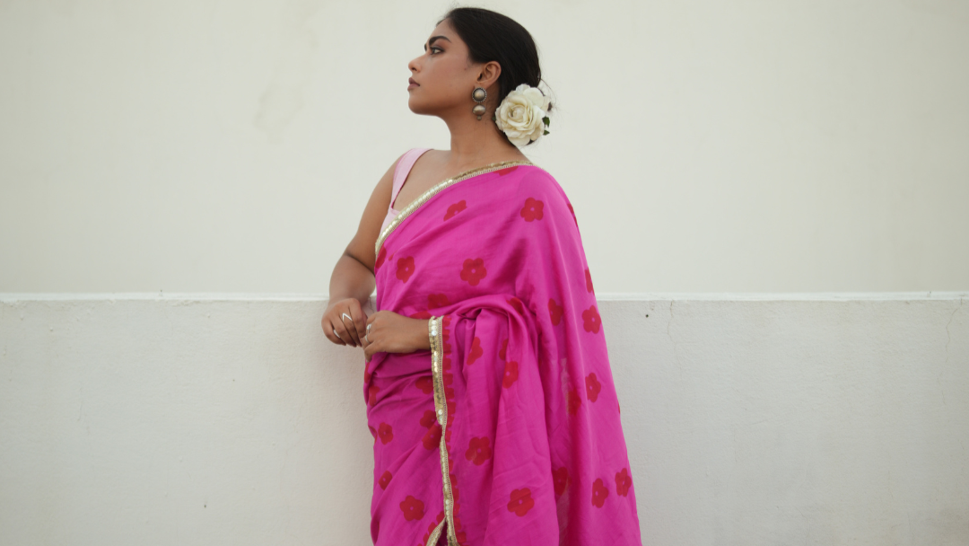 Woman wearing a bright pink saree with red patterns against a white wall.